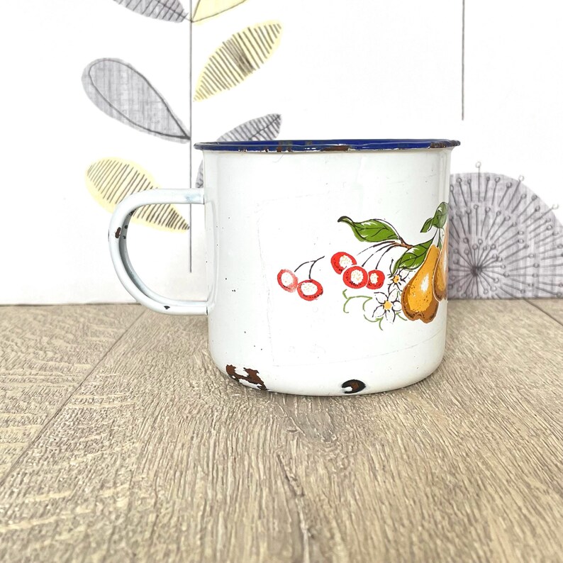 May include: White enamel mug with a blue rim and a floral design featuring pears, cherries, and flowers.