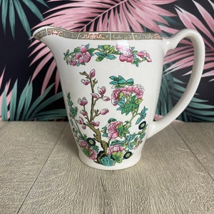 May include: White ceramic pitcher with a floral design featuring pink flowers, green leaves, and brown branches. The pitcher has a curved handle and a decorative border around the rim. The pitcher is a classic piece of kitchenware.