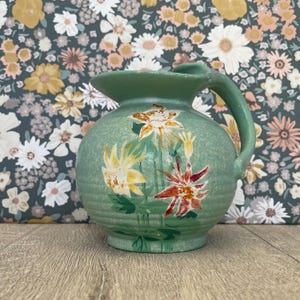 May include: A sage green ceramic pitcher featuring a rounded body with horizontal ribbed detailing and a curved handle. The surface is decorated with hand-painted floral motifs in shades of yellow, white, and deep red, showcasing petals.