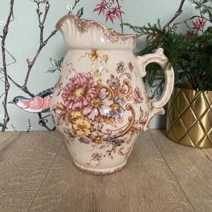 May include: A cream-colored ceramic pitcher featuring a decorative floral pattern in shades of pink, yellow, and blue. The pitcher has a scalloped rim with a brown-toned edge, a curved handle, and intricate scrollwork accents surrounding the flowers.