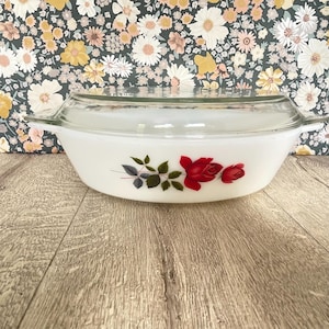 May include: A white oval casserole dish with a clear glass lid. The dish features a floral design of red roses and green leaves. The dish is sitting on a wooden surface.