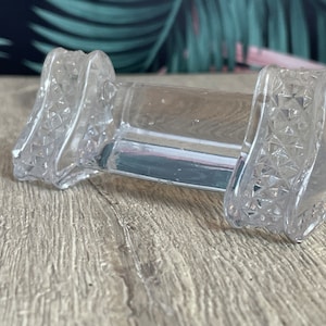 May include: Clear glass napkin holder with diamond-patterned ends. The rectangular holder is designed to hold napkins and is made of transparent glass. The ends have a decorative, textured design. The holder is on a wooden surface.