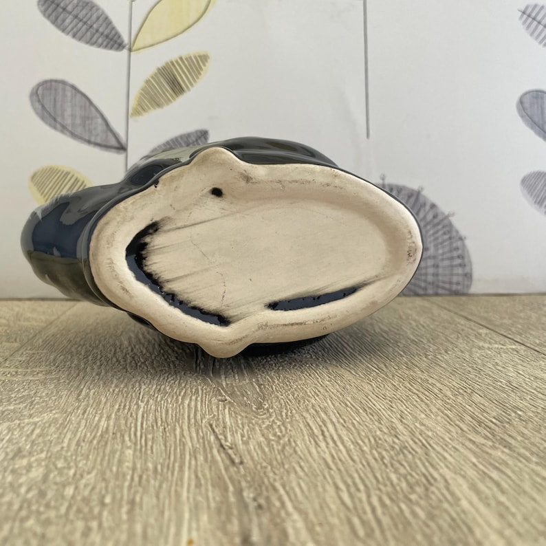 May include: A black ceramic vase with a cream-colored interior. The vase has a unique shape and a glossy finish. The bottom of the vase is visible, showing its oval shape and the texture of the ceramic.
