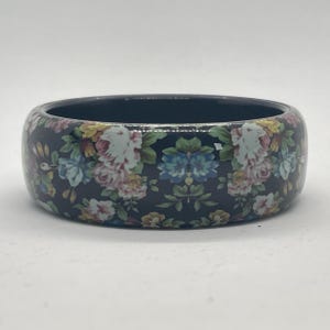 May include: A wide, dark-coloured bangle bracelet with a floral pattern. The bracelet features a dark background with a repeating pattern of pink, blue, yellow, and white flowers. The bracelet is approximately 2.5 cm wide.