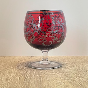 以下が含まれることがあります： 銀色の花の模様が施された赤いガラスのゴブレット。ゴブレットは丸いボウルと円形のベースを持つステムを持っています。