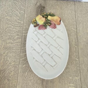 May include: White ceramic oval serving dish with a woven basket design. The top edge features a decorative arrangement of yellow pears, peaches, and red strawberries with green leaves. A charming piece for serving fruit or snacks.
