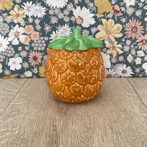 May include: A ceramic pineapple-shaped jar with a green lid. The jar is a warm orange colour with a textured surface, and the lid is green with a stem detail. The jar is on a wooden surface.
