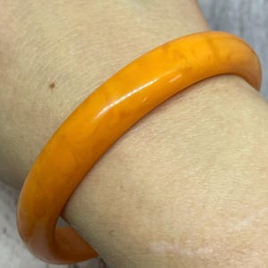 May include: A close-up of a vibrant orange bangle bracelet. The bracelet has a smooth, glossy finish and a marbled appearance. The jewelry is a solid, circular shape, and appears to be made of a hard material.