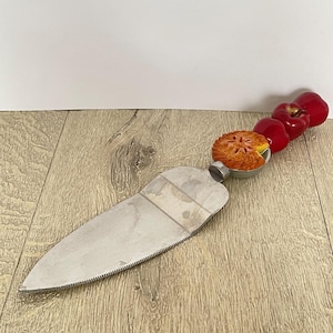 May include: A silver cake server with a serrated edge and a red apple and pie themed handle.