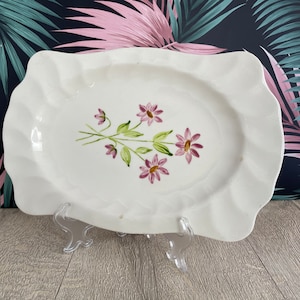 May include: White ceramic serving platter with a ruffled edge and a floral design. The platter features a bouquet of pink flowers with green stems and leaves. The platter is rectangular with a slightly oval centre.