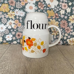 Könnte beinhalten: Weißer Keramik-Mehlbehälter mit Griff und Deckel. Das Wort "flour" ist schwarz aufgedruckt. Der Behälter ist mit orangefarbenen, gelben und braunen floralen Designs verziert.