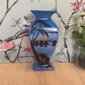 以下が含まれることがあります： 黒と金の風景が手描きされた青い陶器の花瓶。シルエットの木々、金色の屋根を持つ小さな建物、水上のボートが描かれています。花瓶は縁と底が広がった形状をしています。