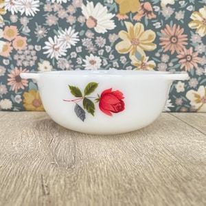 May include: A white, vintage Pyrex bowl with a red rose floral design. The bowl has two handles and is set against a floral patterned background. The rose is detailed with green leaves and a red bloom.