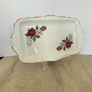 Peut inclure: Un plateau de service rectangulaire blanc cassé avec une bordure dorée et des bords festonnés. Le plateau présente un motif floral de roses rouges et de feuilles vertes. Le plateau est soutenu par un support en plastique transparent.