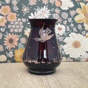 May include: A dark purple glass vase with a flared top and a round base. The vase features a silver painted bird in flight and silver painted grass. The vase is approximately 15 cm tall.