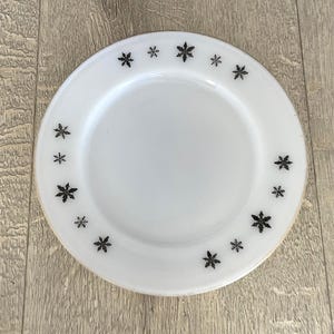 May include: Round white plate with a gold rim, decorated with black snowflake motifs around the edge. A simple, elegant design for serving food. The plate is a classic piece.