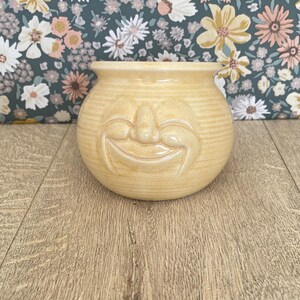 以下が含まれることがあります： 笑顔の顔のデザインが施された、明るい黄色のセラミックポット。 ポットは丸い形で、表面はざらざらしています。 背景は、濃いティール色の背景に花柄が描かれています。 ポットは木製の表面に置かれています。