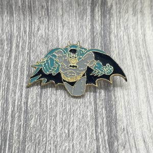 May include: A metal enamel pin featuring a stylised figure in a grey and teal costume with a black cape, depicted in a dynamic forward-reaching pose with gold-toned outlines defining the character's muscular form and pointed cowl mask details.