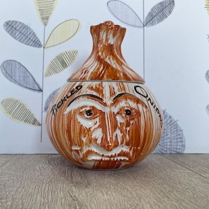 Könnte beinhalten: Ein zwiebelförmiges Keramikgefäß mit Deckel und Gesichtsdesign. Das Gefäß ist hauptsächlich braun mit weißen Akzenten und den Worten "Tickled Onion" um das Gesicht. Der Deckel ist wie die Spitze einer Zwiebel geformt.
