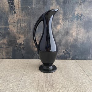 Könnte beinhalten: Ein hoher, glänzend schwarzer Keramikkrug mit einer glatten, geschwungenen Silhouette. Das Gefäß verfügt über einen schlanken Hals, einen breiten, abgerundeten Körper und einen hohen, bogenförmigen Henkel, der vom oberen Hals zur Basis führt.