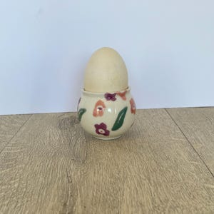 May include: A cream-coloured ceramic egg cup decorated with red, orange, and green floral patterns. It holds a light-coloured wooden egg. The egg cup is on a wooden surface.