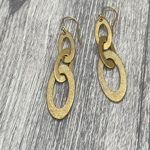 May include: Gold-coloured, linked oval earrings. Each earring has three connected ovals, with the bottom oval having a textured, sandblasted appearance. The earrings hang from a simple hook.