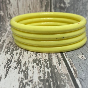 May include: A stack of four pale yellow bangles. The bracelets are made of a smooth, solid material and have a rounded, tubular shape. The bangles are stacked on top of each other, creating a layered effect. The background is a weathered, grey and white wooden surface.