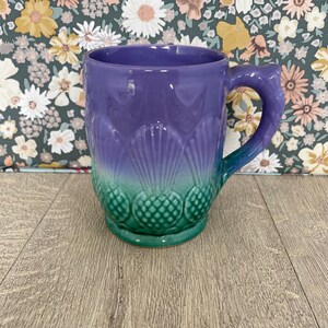 May include: A ceramic mug with a gradient design, transitioning from purple to teal green. The mug features a textured pattern resembling pineapples at the base and a curved handle. The mug is set against a floral patterned background.