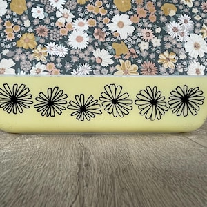 May include: A rectangular yellow baking dish featuring a repeating pattern of six black line-drawn daisy flowers across the front side. The dish has integrated handles on both ends and a smooth, glossy finish, showcasing a simple and functional design.