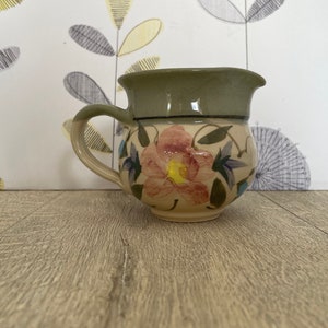 May include: A small, cream-colored ceramic pitcher with a green rim and a floral design. The pitcher has a handle and is decorated with a pink flower and green leaves.