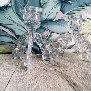 May include: Two clear, faceted crystal candle holders. Each has a square base, an arched center, and a top designed to hold a candle. The candle holders are on a wooden surface with a floral patterned background.