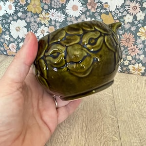 May include: A small, dark green ceramic bowl with a textured, floral-like pattern. The bowl is held in a hand, showcasing its glossy finish and unique design. The bowl's color is a deep, rich green, and it has a small spout.