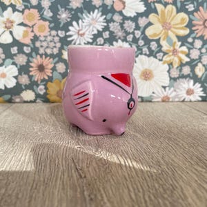 May include: A pink ceramic owl-shaped egg cup. The owl has black and red accents, including a red hat and black lines. The egg cup is on a wooden surface with a floral patterned background.