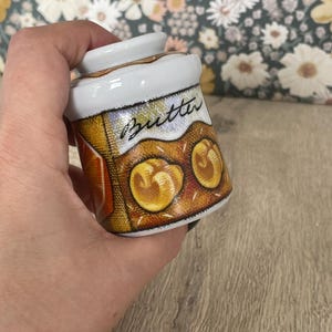 May include: A small, white ceramic butter dish with a lid. The dish features a colorful illustration of butter and rolls, with the word "Butter" written on the front. The dish is held against a floral background.