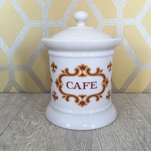 May include: A white ceramic canister with a lid, featuring a decorative design with the word "CAFE" in a red and gold frame. The canister has a bulbous base and a small knob on top of the lid.