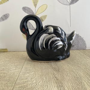 May include: A glossy black ceramic swan figurine with white and silver painted details. The swan has a curved neck and a red accent on its beak. The figurine is on a wooden surface.