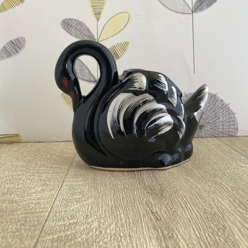 May include: A glossy black ceramic swan figurine with white and silver painted details. The swan has a curved neck and a red accent on its beak. The figurine is on a wooden surface.