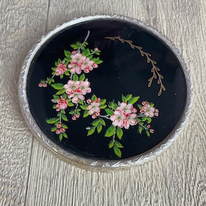 Könnte beinhalten: Runder, antiker Kompakt-Make-up-Spiegel mit schwarzem Hintergrund und floralem Design. Die Blumen sind rosa mit grünen Blättern, und der Spiegel hat einen silberfarbenen, verzierten Rand. Eine goldfarbene, geschwungene Linie ist ebenfalls vorhanden.