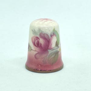 May include: A ceramic thimble with a floral design. The thimble features a pink rose with green leaves against a white background. The base of the thimble is a darker shade of pink. The thimble is approximately 2.5 cm tall.
