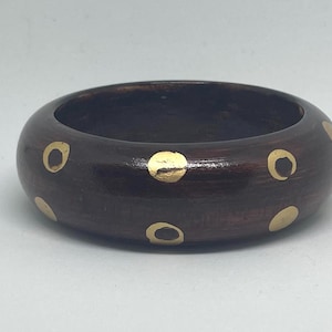 May include: A dark brown wooden bangle bracelet with gold-colored circular accents. The bracelet is thick and has a smooth, polished finish. The gold accents are evenly spaced around the exterior of the bracelet.