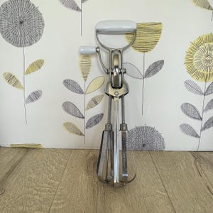 May include: A vintage hand-crank egg whisk with a white handle and a silver metal body. The whisk has a D-shaped handle and a small side handle. The beaters are made of thin metal rods. The background is a floral wallpaper.