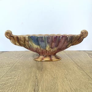 Könnte beinhalten: Eine dekorative Keramikschale mit Sockel und geschwungenen Griffen. Die Schale hat eine Farbpalette aus Braun, Gelb, Blau und Lila. Der Rand hat einen gewellten Rand. Die Schale steht auf einer Holzoberfläche.