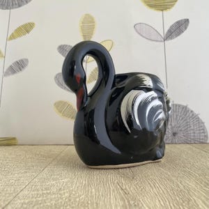 May include: A black ceramic swan-shaped planter with a glossy finish. The swan has white accents on its head and a red beak. The planter is designed to sit on a flat surface and is suitable for small plants or decorative use.