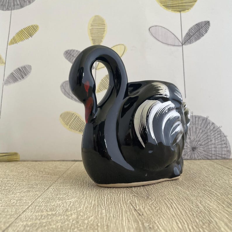 May include: A black ceramic swan-shaped planter with a glossy finish. The swan has white accents on its head and a red beak. The planter is designed to sit on a flat surface and is suitable for small plants or decorative use.