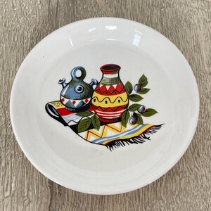 May include: White ceramic plate featuring a vibrant illustration of two vases and foliage. One vase is blue, the other is yellow and red. The plate has a decorative design, suitable for serving or display.