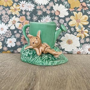 May include: A green ceramic watering can planter featuring a sculpted tan figure of a small creature with pointed ears resting on a textured green base. The watering can has a curved handle and a spout, with the figure positioned in the front.