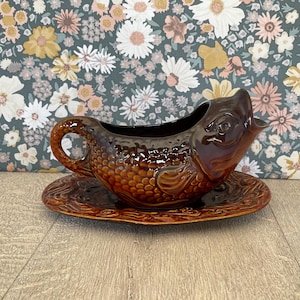 May include: A dark brown ceramic gravy boat shaped like a fish with a textured scale pattern, a curved handle, and an open mouth spout. The gravy boat rests on a matching oval-shaped ceramic underplate with a decorative embossed border design.