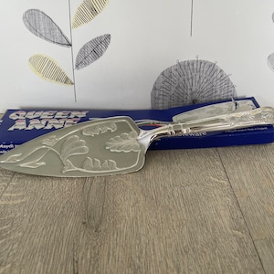 May include: A silver cake server with an engraved floral pattern. The server is on a blue box with the words "QUEEN ANNE" printed on it. The server is made in England.