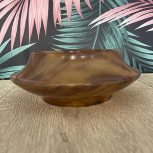 May include: A brown, decorative bowl with a flared rim. The bowl has a glossy finish and a marbled appearance. The bowl is sitting on a wooden surface. The background features a tropical leaf pattern in pink and green.
