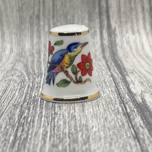 May include: A white porcelain thimble with a colorful bird and floral design. The bird is blue, yellow, and orange, perched on a branch with red flowers. The thimble has gold trim around the top and bottom.